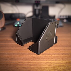 May include: A black desk organizer with multiple compartments and a stylized emblem on the side. The organizer is made of a matte material and sits on a wooden desk. The design is angular and modern.