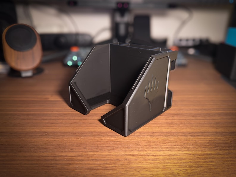 May include: A black desk organizer with multiple compartments and a stylized emblem on the side. The organizer is made of a matte material and sits on a wooden desk. The design is angular and modern.