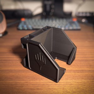 May include: A black desk organizer with multiple compartments and a stylized emblem on the front. The organizer is made of a matte material and sits on a wooden surface. The design includes angled sides and a front opening.