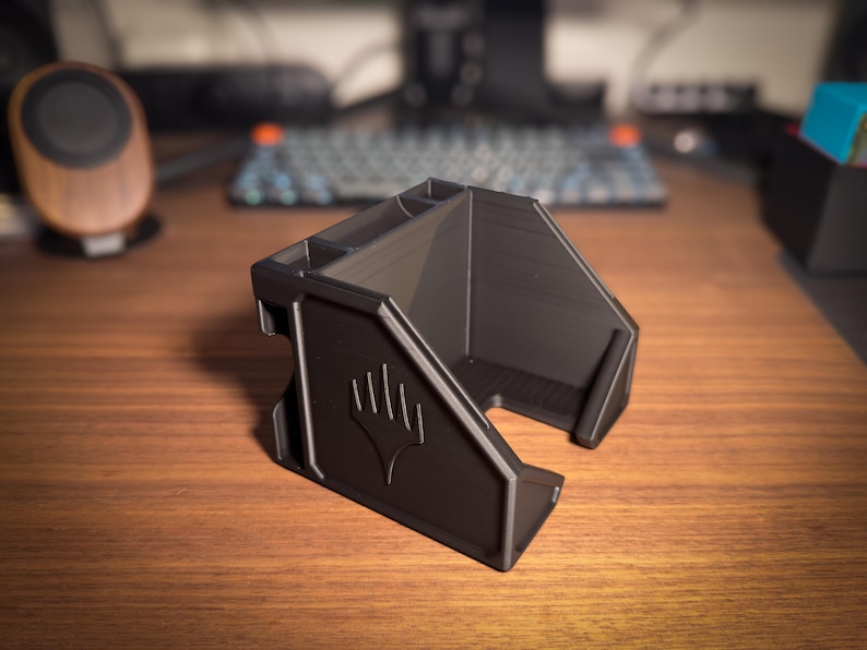 May include: A black desk organizer with multiple compartments and a stylized emblem on the front. The organizer is made of a matte material and sits on a wooden surface. The design includes angled sides and a front opening.