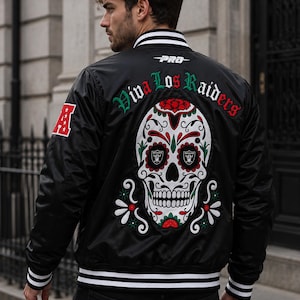 Pode incluir: Uma jaqueta universitária de cetim preto com uma grande caveira bordada nas costas em tons de vermelho, branco e verde. O texto "Viva Los Raiders" está arqueado acima da caveira, com um emblema vermelho "A" na manga direita e listras brancas.