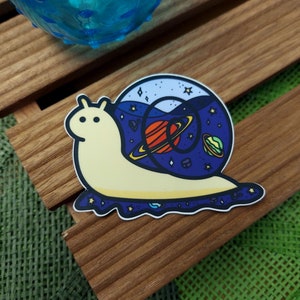 May include: A yellow snail sticker with a blue shell filled with stars, planets, and a galaxy. The snail is on a wooden surface.
