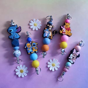 May include: Several beaded keyrings with cartoon character charms, including Stitch, Winnie the Pooh, and Minnie Mouse. Each keyring has a silver ring and colourful beads. White daisy decorations are scattered around the keyrings.