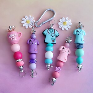 May include: A selection of beaded keyrings with medical motifs. Each keyring has a charm shaped like a nurse's uniform, a coffee cup with the text "NURSE LIFE", or a nurse figurine. The beads are in shades of pink, purple, blue, and white.