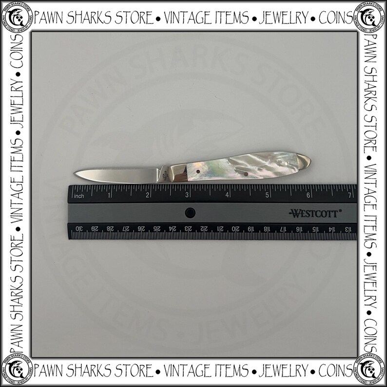 May include: A folding pocket knife with a metallic blade and a pearlescent handle, positioned above a black Westcott ruler. The knife measures approximately 3.5 inches in length when closed, as indicated by the visible imperial measurement markings.