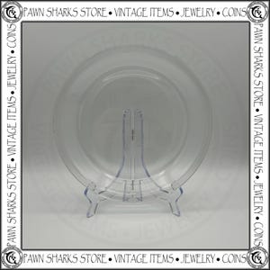May include: A clear, circular glass plate displayed on a transparent plastic easel stand. The plate features a smooth, reflective surface with a simple, unadorned rim. The stand is designed with two front-facing supports to hold the plate upright.