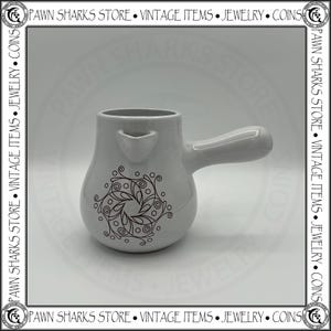 May include: A white ceramic pitcher with a single handle and a small pouring spout. The body of the pitcher features a black printed graphic design consisting of swirling lines, leaf-like shapes, and small circles arranged in a circular pattern.