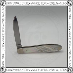 May include: A folding pocket knife with a single silver-toned blade and a handle featuring a pearlescent white finish. The base of the blade is stamped with the text USA TB61028 SS. The knife is displayed in an open position against a neutral background.