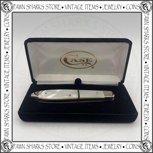 May include: A closed folding pocket knife with a white pearlescent handle and metallic bolsters, resting inside a black velvet-lined display case. The interior lid features the gold-colored text CASE XX. The case is framed by a border with text.