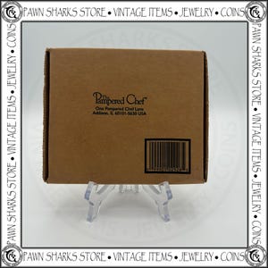 K&ouml;nnte beinhalten: Ein brauner Pappkarton mit dem Text "The Pampered Chef" und Adressinformationen. Ein Barcode ist unten rechts sichtbar. Die Box wird auf einem transparenten Kunststoffst&auml;nder pr&auml;sentiert.