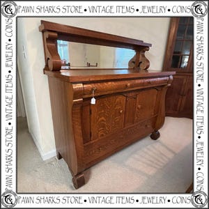 May include: A vintage wooden sideboard with a mirror, featuring a warm brown finish. The cabinet has two doors, drawers, and decorative carvings. The piece is a classic example of antique furniture.