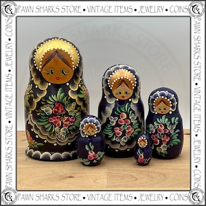 May include: A set of five dark blue nesting dolls featuring hand-painted floral patterns in red, green, and white. Each wooden doll depicts a figure with a gold-toned head covering, detailed facial features, and decorative scalloped borders.