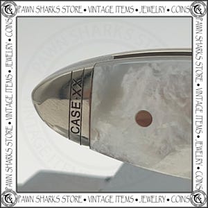 May include: A close-up view of a pocket knife handle featuring a polished silver metal bolster engraved with the text CASE XX. The handle has a white, marbled, pearlescent finish with a single circular rivet visible on the side of the grip.