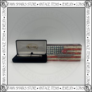 May include: A W.R. Case & Sons Cutlery Co. pocket knife with a white handle rests inside a black velvet-lined display case. Beside it sits a rectangular cardboard box featuring a distressed American flag print and the text W.R. CASE & SONS CUTLERY CO.