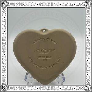 K&ouml;nnte beinhalten: Ein herzf&ouml;rmiger Steinzeug-Schmuckanh&auml;nger von The Pampered Chef mit dem Text "Peace on Earth Heart" und "Family Heritage Stoneware". Der Anh&auml;nger ist hellbraun und hat ein Loch zum Aufh&auml;ngen. Das Jahr 2002 ist ebenfalls sichtbar.