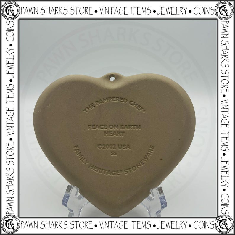 K&ouml;nnte beinhalten: Ein herzf&ouml;rmiger Steinzeug-Schmuckanh&auml;nger von The Pampered Chef mit dem Text "Peace on Earth Heart" und "Family Heritage Stoneware". Der Anh&auml;nger ist hellbraun und hat ein Loch zum Aufh&auml;ngen. Das Jahr 2002 ist ebenfalls sichtbar.