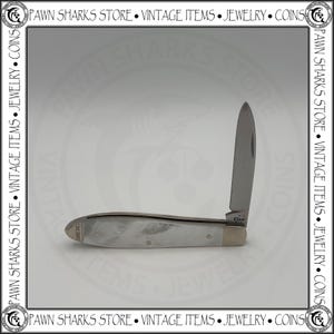 May include: A folding pocket knife with a single silver-toned blade and a handle featuring a pearlescent white marbled finish. The handle includes a metallic bolster and a small metal inlay at the end marked with the text XX 1889.