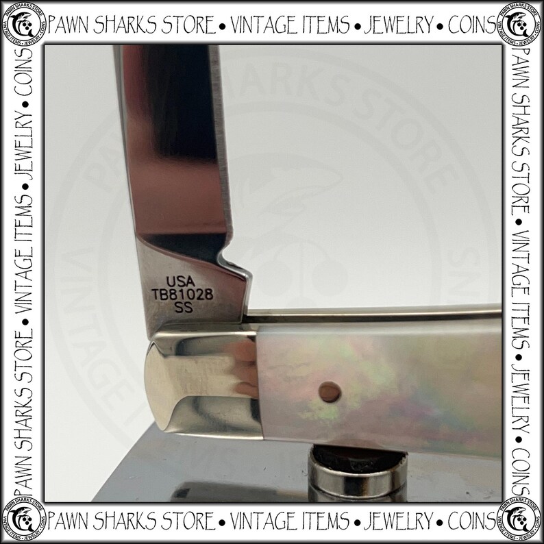 May include: A folding pocket knife with a polished metal blade and a handle featuring an iridescent, pearlescent finish. The blade is stamped with the text USA, TB81028, and SS near the bolster. The knife has a silver-toned metal bolster.