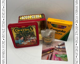 May include: A vintage Crayola crayon set, featuring a red metal box with a 1992 Crayola design, an open yellow box of 64 crayons, and a small container of crayons. Also included is a promotional insert.