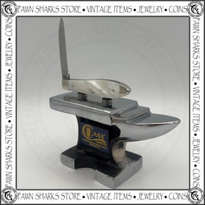 May include: A silver-colored metal anvil display stand featuring a Case Tested XX logo label on the front. Resting on top of the anvil is a folding pocket knife with a white pearlescent handle and a single upright silver blade.