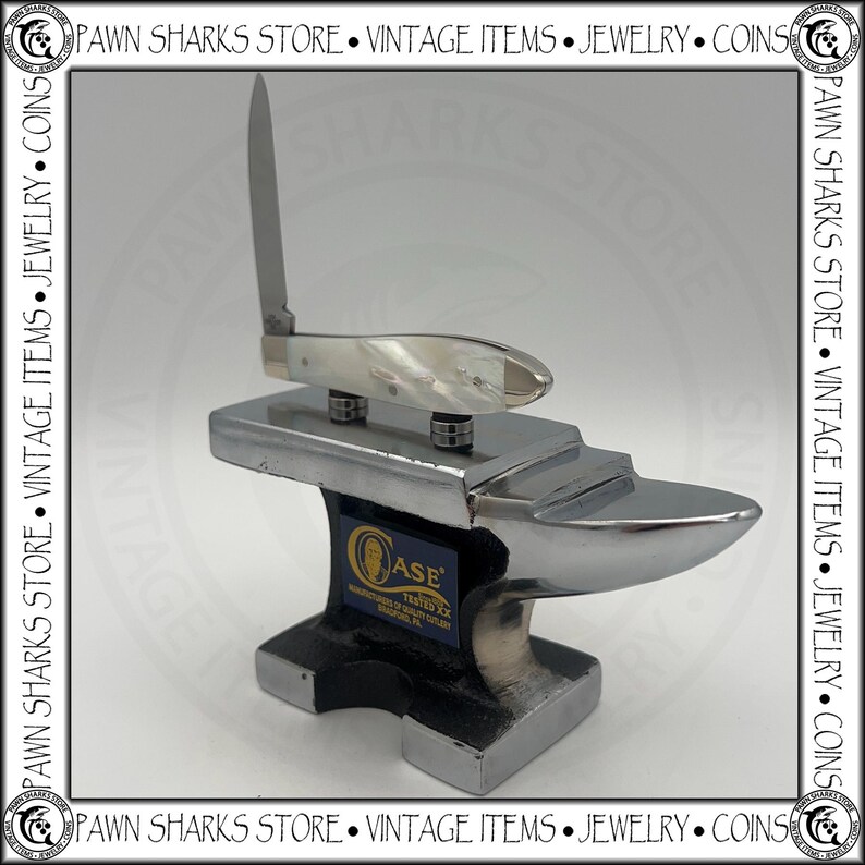 May include: A silver-colored metal anvil display stand featuring a Case Tested XX logo label on the front. Resting on top of the anvil is a folding pocket knife with a white pearlescent handle and a single upright silver blade.