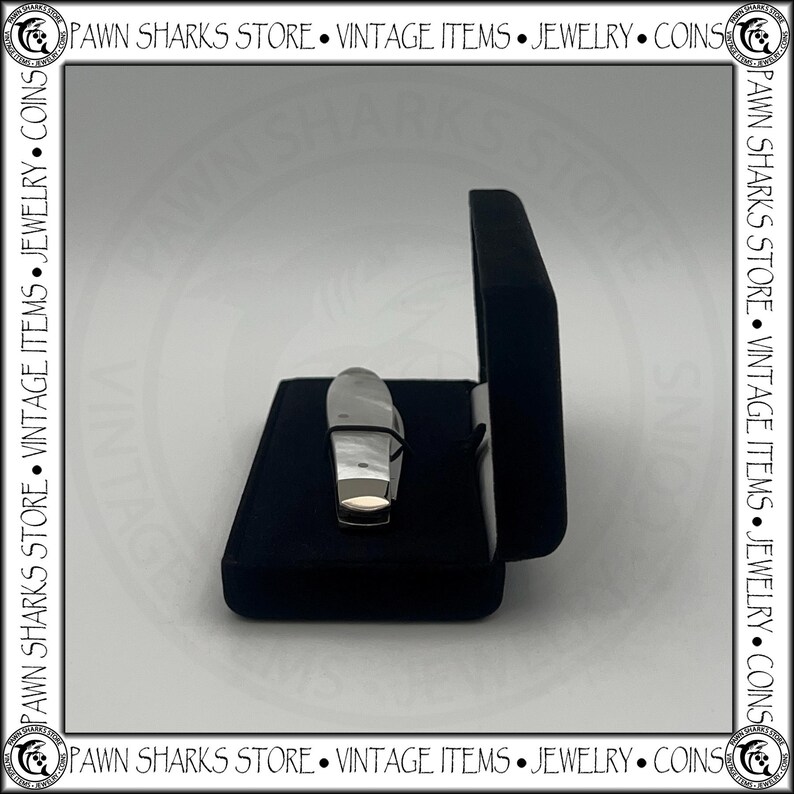 May include: A small folding pocket knife with a smooth, light-colored handle and a metallic bolster, resting inside a dark blue velvet-lined display case. The case is open, revealing the knife positioned horizontally against the dark fabric interior.