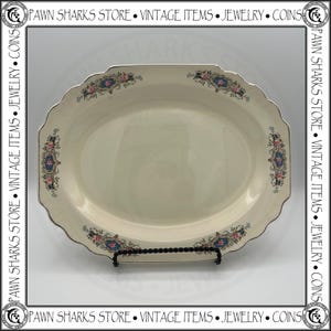 May include: An oval-shaped cream-colored serving platter with a scalloped edge and a silver-toned rim. The border features four symmetrical floral motifs consisting of pink roses, blue accents, and black scrollwork details on a white background.