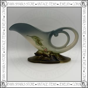 May include: A ceramic gravy boat featuring a gradient of slate blue to cream, accented with a sculpted green leaf and small pink flower motif at the base. The vessel has a curved handle and a wide pouring spout, finished with a dark brown base.