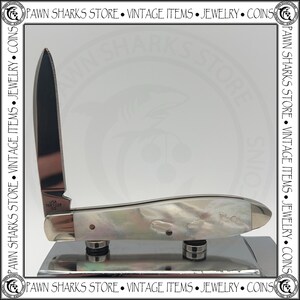 May include: A folding pocket knife with a single upright silver-toned blade and a handle featuring an iridescent mother-of-pearl finish. The blade is stamped with the text USA T681028 SS. The knife is positioned on a reflective metallic base.