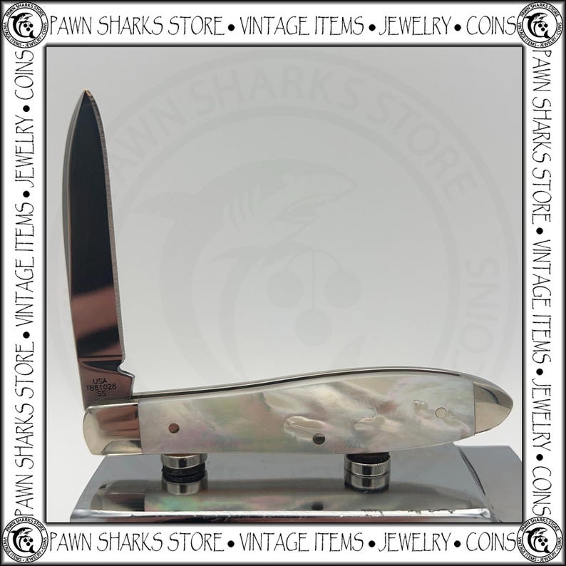 May include: A folding pocket knife with a single upright silver-toned blade and a handle featuring an iridescent mother-of-pearl finish. The blade is stamped with the text USA T681028 SS. The knife is positioned on a reflective metallic base.