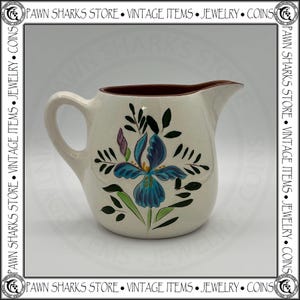 May include: A cream-colored ceramic creamer pitcher featuring a hand-painted blue iris flower design with green leaves and black accents. The pitcher has a curved handle, a pouring spout, and a dark reddish-brown interior rim finish.