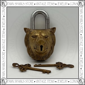 May include: A brass padlock shaped like a lion's head with a silver-toned metal shackle. The keyhole is located at the lion's mouth. Two matching brass keys with ornate, decorative handles are positioned horizontally in front of the padlock.