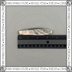 May include: A folding pocket knife with an iridescent mother-of-pearl handle and silver-toned metal bolsters. The knife is positioned above a black ruler, showing a total length of approximately 3.5 inches when in the closed position.