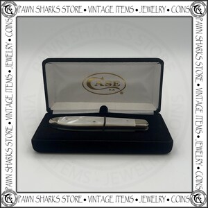 May include: A closed folding pocket knife with a white pearlescent handle and silver metal bolsters, resting inside a black velvet-lined display case. The interior lid of the case features the gold-colored Case XX logo on a white fabric background.