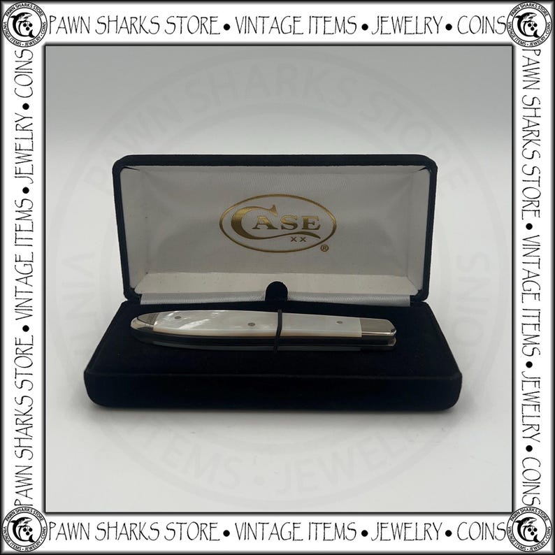May include: A closed folding pocket knife with a white pearlescent handle and silver metal bolsters, resting inside a black velvet-lined display case. The interior lid of the case features the gold-colored Case XX logo on a white fabric background.