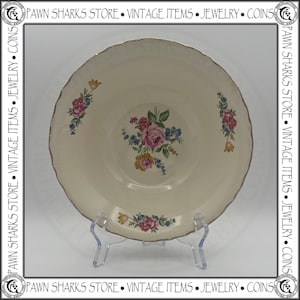 May include: A round, cream-colored ceramic bowl with a scalloped edge and a metallic gold rim. The center features a floral bouquet with pink, yellow, and blue flowers and green leaves. Smaller matching floral sprays are positioned on the sides.
