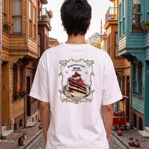 May include: A white short-sleeve t-shirt featuring a graphic on the back. The design shows a slice of chocolate cake with a strawberry on a plate, framed by an ornate border with the text Peretti's Cafe Menu Da Semara and small stars.