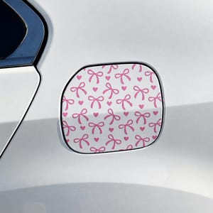 May include: A white car fuel door cover featuring a repeating pattern of small pink bows and pink hearts. The decal is applied to the square-shaped fuel door of a white vehicle, covering the entire surface with the pink and white graphic design.