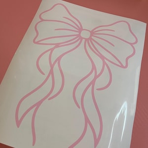 May include: A light pink vinyl decal featuring a line art illustration of a decorative bow with long, flowing ribbons. The design is printed on a white rectangular backing sheet, showcasing clean, smooth lines that form the bow shape.