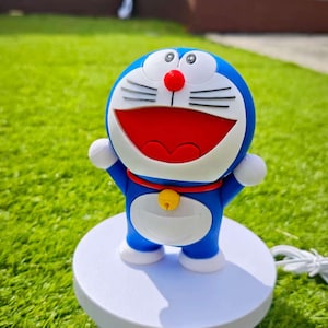 May include: A blue Doraemon figurine with a red mouth, nose, and collar, and white accents. The figure is standing on a white, round platform. The background is a green lawn and a wooden fence.
