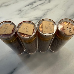 May include: Four clear plastic tubes filled with copper-coloured coins. Each tube is sealed with a clear plastic lid and a small piece of paper with handwritten numbers. The tubes are arranged on a white and grey marble surface.