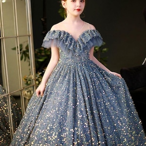 May include: A formal, floor-length gown in a shimmering blue-gray hue. The dress features an off-the-shoulder design with ruffled trim and a full, flowing skirt. The bodice and skirt are covered in sparkling embellishments, and a delicate tiara adorns the head.