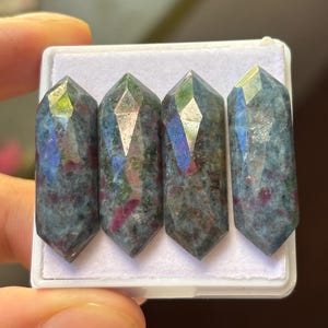 Peut inclure: Quatre baguettes de cristal de rubis en zoïsite facettées. Les baguettes présentent un mélange de teintes vert foncé, bleu et rouge. Elles sont exposées dans un récipient rectangulaire blanc. Les cristaux ont un sommet pointu et un design à facettes.
