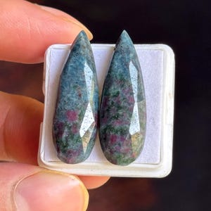 Peut inclure: Deux pierres précieuses facettées en forme de poire présentant un motif tacheté d'inclusions bleu-vert profond et rose magenta vif. Ces pierres polies sont présentées côte à côte sur un petit plateau en plastique blanc avec un insert en papier.