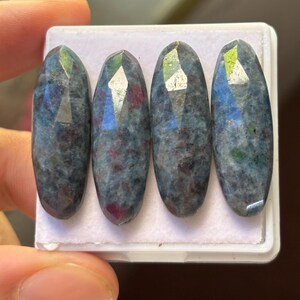 Peut inclure: Quatre pierres précieuses ovales avec des surfaces à facettes. Les pierres ont une couleur de base bleu-gris foncé avec des inclusions rouges et vertes. Elles sont présentées sur une surface blanche.