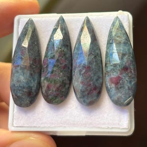 Peut inclure: Quatre pierres précieuses taillées en forme de goutte. Les pierres ont une couleur de base bleu-gris avec des inclusions rouges et vertes. Elles sont présentées dans un plateau blanc, mettant en valeur leurs surfaces polies et leurs motifs uniques. Ces pierres sont adaptées à la création de bijoux.