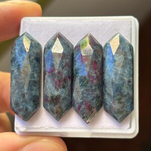 May include: Four faceted, blue-green crystal wands with red inclusions are displayed in a white tray. The crystals have a geometric shape with pointed ends. The wands are held by a person's fingers.