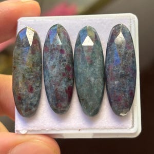 May include: Four oval-shaped gemstones with faceted tops. The stones have a mix of colors, including blue-green and red. They are set in a white tray, and the gemstones are approximately 1 inch in length.