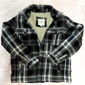Puede incluir: Chaqueta de camisa de franela a cuadros negros, blancos y grises con forro de sherpa color crema. La chaqueta tiene dos bolsillos en el pecho y cierres de bot&oacute;n. La etiqueta dice "AMERICAN EAGLE OUTFITTERS".