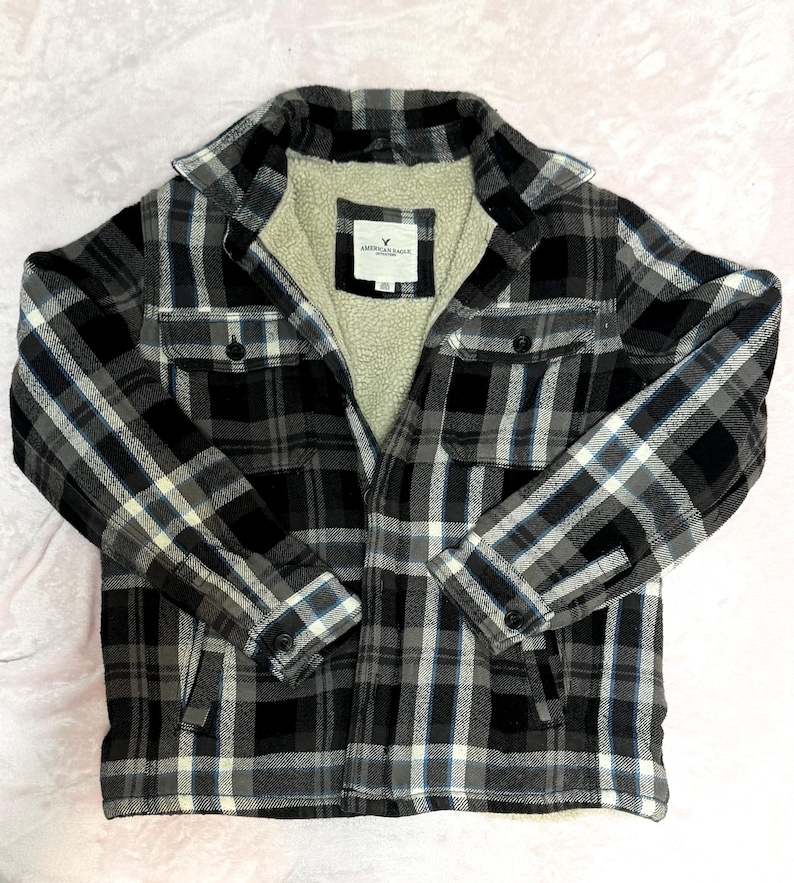 Puede incluir: Chaqueta de camisa de franela a cuadros negros, blancos y grises con forro de sherpa color crema. La chaqueta tiene dos bolsillos en el pecho y cierres de bot&oacute;n. La etiqueta dice "AMERICAN EAGLE OUTFITTERS".